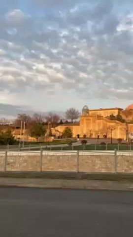 The camera captures a static, mid-range view of a tan-colored building with a distinctive domed roof, likely a mosque or cultural center, set against a backdrop of a cloudy, early-morning or late-afternoon sky. The building is situated on a slightly elevated landscape, separated from the viewer by a stone wall and a narrow road. The light from the sky illuminates the building, enhancing its architectural details. The surrounding area features sparse greenery, including trees and hedges, typical of a well-maintained public park or garden. The scene suggests a calm, tranquil setting, focusing on the visual impact of the building and its surroundings.