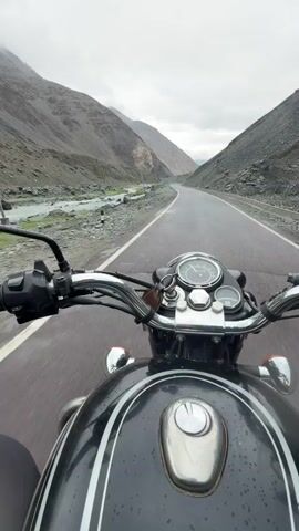 The video begins with a first-person perspective shot from a motorcycle traveling on a paved road. The road is wet, suggesting recent rain. On the left side of the road, a shallow, rocky stream flows alongside. The surrounding environment is mountainous, with steep, barren slopes on either side of the road. The sky is overcast with clouds. The motorcycle is black and chrome, with visible handlebars, mirrors, a speedometer, and part of the fuel tank. The camera's movement captures the feeling of riding the motorcycle through the mountainous landscape.