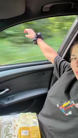 This media file shows two individuals inside a car. The first subject, a woman, is in the passenger seat. She is wearing a black t-shirt with a colorful logo on the front, possibly a stylized "LV" or similar branding. Her eyes are closed, and she appears relaxed or enjoying the moment, with her arm outstretched and head tilted back. Green foliage is visible through the passenger window, suggesting a tree-lined road or rural setting. The second subject, a man, is driving the car. He is wearing a white t-shirt and gray shorts. He has a watch on his left wrist and a bracelet on his right. He is looking forward, focused on driving. The interior of the car appears to be black leather. The scene is captured from inside the car, with the camera focusing alternately on the passenger and the driver. The lighting suggests it is daytime, possibly overcast due to the diffused light and slight blur in the background, indicating motion. The location context provided is Khadzhiysko, Bulgaria. The overall mood seems casual and possibly celebratory or a shared moment of enjoyment during a drive.