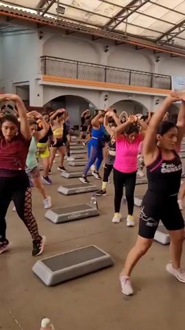 A group of people, primarily women, are engaged in a step aerobics class in a large, open indoor space with a high, arched ceiling and exposed beams. The floor is concrete. Each participant has a personal aerobic step platform in front of them. The class is being led by an instructor at the front of the room, who is not clearly visible in this frame, but their presence is implied by the synchronized movements of the participants. Most of the participants are wearing athletic attire, including leggings and sports tops. The atmosphere appears energetic and focused.