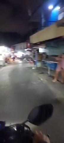 The video shows a street at night, possibly in Southeast Asia, with several people on motorcycles. The street is narrow and lined with buildings, some of which appear to be shops or homes. There are also several motorcycles parked along the side of the road, and some people are sitting on benches or standing. The lighting is dim, with some artificial lights illuminating the scene. The overall impression is one of a bustling nighttime street scene in a residential or commercial area.