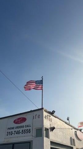 The video shows an American flag waving on a flagpole mounted on the corner of a white building. The building has a sign that says "Perry's Cafe" and provides a phone number for "PHONE ORDERS CALL 310-746-5556". The building also has the numbers "2400" visible. In the background, a clear blue sky is present, with the sun casting a lens flare on the right side of the frame. Another flag, possibly a California state flag, is visible flying on a pole on the right side of the building. The camera appears to be positioned below the flag, looking up at an angle. The video is static with no movement or people visible.