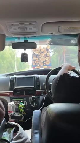 The video shows the interior of a car from the perspective of a passenger. The driver, whose back is to the camera, is steering the vehicle. In front of the car, a truck loaded with logs is visible on the road. The dashboard of the car has a navigation screen, a gear shift, and drink cans on a tray. The environment outside the car appears to be a rural or forested area.