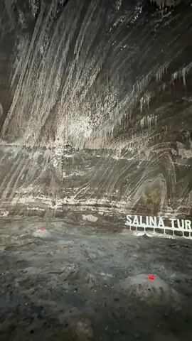 The video opens with a close-up view of the textured, salt-lined ceiling and walls of a cave, with formations resembling icicles hanging from above. The walls exhibit distinct horizontal layers in shades of gray and white. In the foreground, a white sign reads "SALINA TURDA," and several small, red lights are visible on the ground. The camera then pans upward and to the right, revealing a vast underground cavern. In the distance, a large Ferris wheel structure made of metal is illuminated by numerous lights. A suspended walkway with lights also runs along the cavern wall. Several people are visible in the lower part of the frame, walking and looking around the cavern. The overall environment appears to be a tourist attraction within a salt mine.