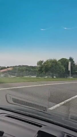 This is a first-person view from inside a vehicle driving on a paved road. The sky is bright blue. Trees and grassy fields line the road. There are streetlights visible in the distance. Reflections on the windshield show some of the interior of the car, including the dashboard and part of the side mirror. There are also some white streaks or smudges on the windshield. The road has a white line marking the edge and a broken white line in the middle. The car appears to be moving at a moderate speed.