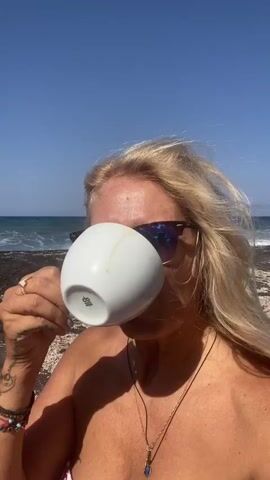 A medium shot shows a fair-skinned woman with blonde hair and sunglasses drinking from a white cup while standing on a beach. The woman is wearing a necklace and appears to be topless, though a portion of a patterned bikini top is visible at the bottom of the frame. Her hand is holding the cup to her lips, and her hair is blowing in the wind. The background shows a body of water with waves and a clear blue sky. The beach appears to be made of dark sand and small shells. The sunglasses reflect the blue sky and the sun. The woman smiles at the camera as she lowers the cup.