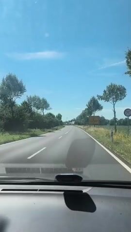 The video shows a first-person perspective from inside a vehicle driving down a paved road. The road is flanked by green trees and vegetation on both sides. The sky is blue with scattered white clouds. There are cars on the road ahead and to the side of the vehicle. There are also road signs visible. The time stamp shows the video is approximately 5 seconds long.