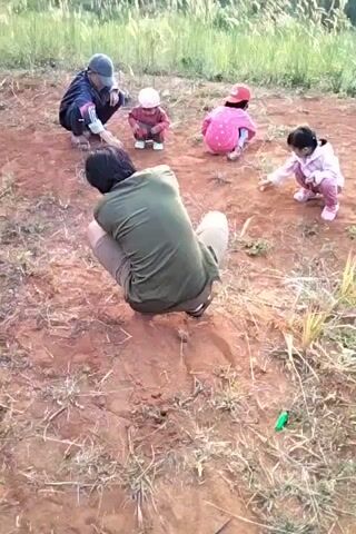 A group of five people, two adults and three children, are crouched on a patch of dry, reddish-brown dirt surrounded by dry grass and vegetation. In the background, a grassy hill rises with trees on top. The adult on the far left is wearing a dark jacket and a baseball cap. Next to them is a child in a pink outfit. The third person from the left is a child in a pink polka dot shirt and red hat. To their right is another child in a light pink outfit. The fifth person, an adult, is in the foreground, facing away from the camera, wearing a green shirt and brown pants. They all appear to be engaged in some activity on the ground, possibly playing with small objects. The lighting suggests it is daytime, with soft, natural light.