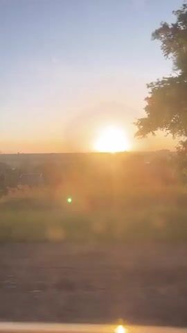 The video shows a view from a moving vehicle, possibly a car or train. The sun is setting or rising on the horizon, creating a bright glow. In the foreground, there is grassy land with some trees and a few buildings. The sky is a gradient of blue to orange, indicating the sunrise or sunset. The camera moves past trees and the landscape, creating a blurred effect.