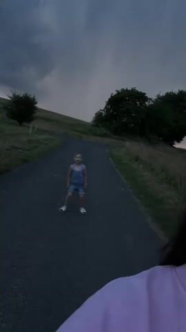 A young boy in a blue tank top and denim shorts stands on a paved path. He is smiling and looking at the camera. The path curves up a grassy hill with trees on either side. The sky is dark and cloudy, with a hint of rain in the distance. The footage is shaky, suggesting it was filmed handheld.