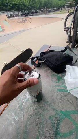 A hand with a ring opens a can of beer, causing it to foam. The scene appears to be a skatepark, with a skateboard and a bag on a concrete surface in the foreground. In the background, there is a skate ramp and a bicycle. The overall lighting suggests it is daytime.
