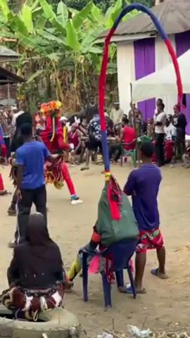 The video shows a group of people dancing in what appears to be an outdoor event or celebration. Several of the dancers are dressed in colorful, traditional attire, including elaborate headgear and costumes with fringed elements. Other individuals in the vicinity are dressed more casually, and some are sitting and observing the dance. The background includes a small building, trees, and a white tent with purple curtains. Overall, the scene seems to capture a cultural performance or festive gathering taking place outdoors.