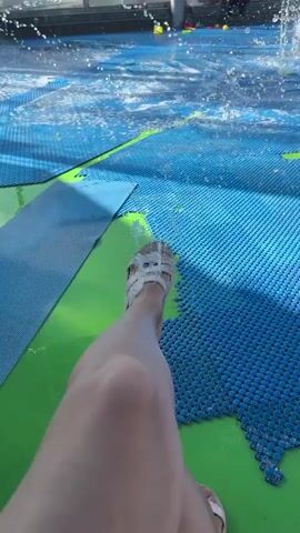 The video shows a person's leg and foot wearing a white sandal, resting on a blue mat with green accents in a splash pad area. Water is spraying from various features, including rings and ground jets, creating a wet surface. In the background, other people are visible, some playing in the water. The scene appears to be outdoors on a sunny day, with a building and canopy structures in the distance.