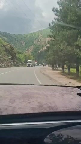 The video shows a car driving along a highway. There is a large hill to the right of the road. There are cars and a truck on the road. There is a man standing on the side of the road. There are three flags and a speed limit sign next to the road. The sky is cloudy.