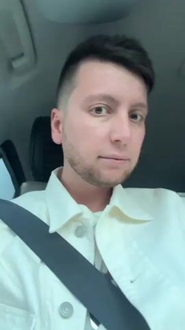 A person is sitting in the driver's seat of a car, wearing a seatbelt and looking directly at the camera. The person is wearing a light-colored button-down shirt and a necklace.  A car window and building can be seen in the background. The car appears to be in motion, as suggested by the changing background. The person looks up towards the top of the car. The audio suggests music is playing.