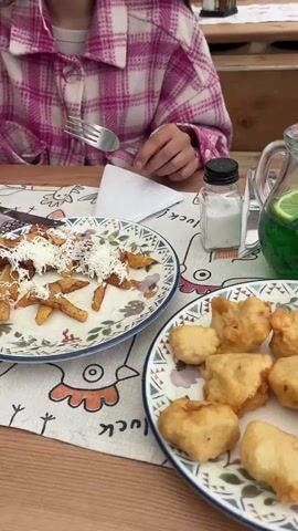 The video showcases two women enjoying a meal at an outdoor restaurant with a scenic view of mountains and a building with a red tile roof. The initial shot displays plates of food, including what appears to be fried fish, a dish with shredded cheese and fried triangles, and a pitcher of green liquid with lime slices. One woman, wearing a pink plaid jacket, is seen with a fork in her hand. The scene shifts to a different table where another woman in a grey hoodie is shown spreading food, possibly a fried item, on a piece of bread or flatbread. A bowl of food with sausage slices is also visible on this table. The video concludes with a pan up to the landscape showing the mountains, the building with a red roof, and various trees under a partly cloudy sky.
