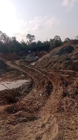 A construction site is shown under a bright, possibly sunny sky. The foreground is dominated by piles of brown dirt and gravel, with tire tracks indicating vehicle activity. A partially paved area, possibly a road or foundation, is visible. In the middle ground, several excavators are operating, digging and moving earth. Trees and vegetation line the background, suggesting a rural or semi-rural location. A white plastic bag, attached to a long pole, is prominent in the center of the frame, appearing to be a marker or a windsock. The overall impression is one of ongoing construction or land development.