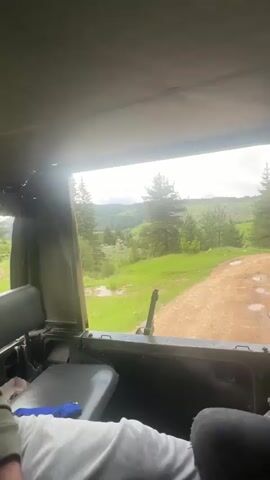 From inside the back of a military vehicle, looking out the window, the camera shows a dirt road in the countryside. The road leads to green fields and trees, with mountains visible in the background. Two people are seated in the back of the vehicle. One is wearing a green hoodie and a wristwatch. The other is wearing a white shirt and dark pants. The vehicle is moving along the road.