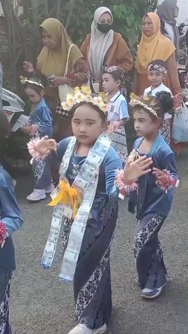 The video shows a group of children, primarily girls, dressed in traditional attire, possibly for a cultural performance or ceremony. They are adorned with floral headpieces and layered clothing. One prominent girl in the foreground wears a blue dress with a decorative sash made of what appears to be money. She is also wearing arm decorations. Another girl beside her, also in blue, is similarly dressed. In the background, other children and adults are visible, some wearing hijabs, suggesting a cultural event. The setting appears to be outdoors, with a paved surface beneath the children's feet. The children seem to be engaged in some form of movement, possibly dancing. The overall atmosphere appears festive and celebratory.