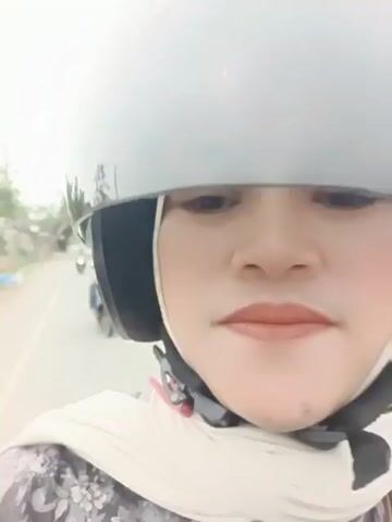 A close-up shot shows a woman wearing a gray helmet and a light-colored hijab with a floral print scarf. Her face is partially visible, with makeup on her lips. She is riding on a motorcycle, as indicated by the blurred background of a road with trees and other motorcycles in the distance. The video appears to be taken from the perspective of the rider.