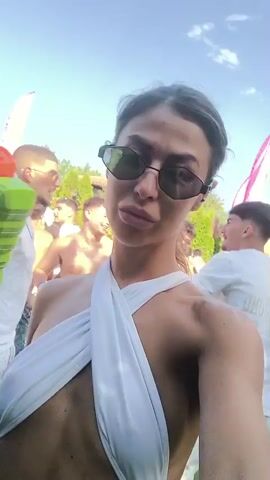 The video shows a woman in a white swimsuit holding a water gun. She is wearing sunglasses and has her hair pulled back. She is taking a selfie in a crowded outdoor setting. There are other people in the background, many of whom are also wearing white. The environment appears to be a party or festival with flags and tents visible. The lighting suggests it is daytime.