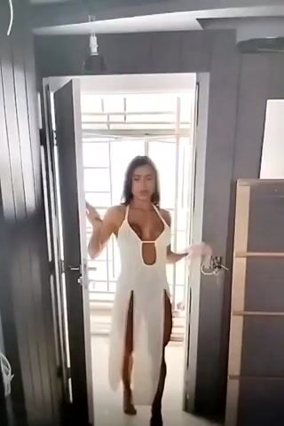 The media file shows a woman in a white dress with high slits walking through a doorway. She has long dark hair and is wearing a halter-neck dress that reveals her chest and legs. The background appears to be an indoor setting with a doorway that opens to a bright area, possibly outdoors or a well-lit room. There are some construction materials or furniture visible in the background. The lighting suggests it could be daytime. The woman's expression is lively, and she appears to be in motion.