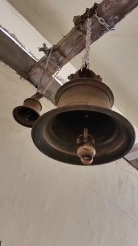 A hand reaches out and rings a large brass bell hanging from wooden beams. The bell has a clapper inside. The scene then shifts to an outdoor courtyard with a white building in the background, possibly a temple or religious site. Several people are gathered in the courtyard, some taking photos with their phones. A mountainous landscape is visible behind the building. The sky is cloudy.