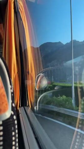 The video is taken from the perspective of someone inside a bus, looking out the window. The window has orange curtains on the left side and a reflection of a side mirror with a round orange indicator light is visible. Outside, there is a clear blue sky and a large mountain range in the background. In the middle ground, a road curves to the right, bordered by green grass and trees. There are some buildings and structures visible in the distance. The bus appears to be moving, as evidenced by the slight blur of the landscape.