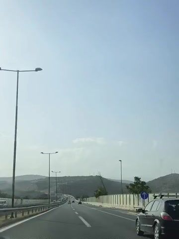The video shows a first-person perspective of driving on a highway. The sky is clear and blue, with a few scattered clouds. The road is paved and has multiple lanes. There are streetlights and guardrails along the side of the road. In the distance, there are rolling hills covered in vegetation. Other vehicles can be seen on the highway, moving in the same direction. The video appears to be filmed during the daytime, possibly late afternoon due to the lighting conditions.