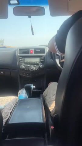 The video begins inside a car, focusing on the dashboard and a person's hands on the steering wheel. The camera then pans to the right, showing a glimpse of the car's interior and the driver. The scene shifts to the view outside the car, revealing a rural landscape with houses and trees under a clear sky. The video continues to show the view from inside the car, looking out the side window, which has a reddish tint, obscuring the details of the passing scenery. The shot then moves to the back seat, focusing on a car door and a portion of the seat.