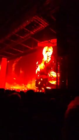 The video shows a concert scene, dominated by red lighting and a large screen displaying a performer. The stage setup is complex, with a visible lighting rig and multiple screens. A crowd is visible in the foreground, silhouetted against the bright stage lights. The main focus is on the screen displaying the singer, who appears to be in the middle of a performance. The singer is wearing a shimmering top. A laser beam can be seen in the upper right of the frame.