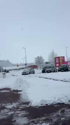 The video shows a snowy scene with a parking lot covered in snow, several cars parked, a large truck on the left, and a KFC sign in the background. Snow is falling heavily. The ground is a mix of red brick and snow-covered asphalt. There are lamp posts and road signs visible. The overall environment suggests a rest stop or roadside service area during a snowstorm.