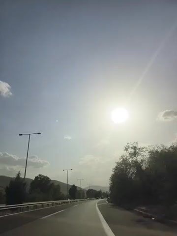 The video shows a road in a rural area with trees and hills on the side. The sun is visible in the sky, casting a bright light. The road appears to be a highway with multiple lanes. There are streetlights on the side of the road and some cars can be seen in the distance. The weather appears to be clear and sunny.
