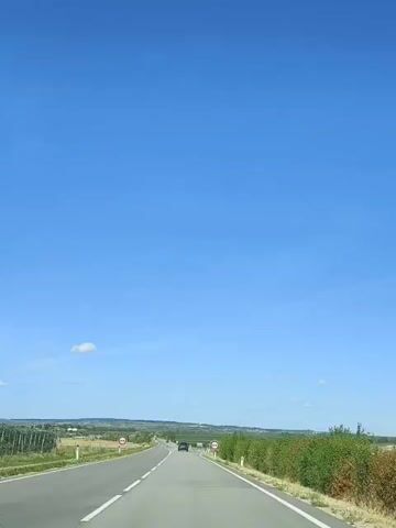 The video shows a clear blue sky and a road with farmland on either side. There are several traffic signs along the road. In the distance, there are a few cars and some trees. The camera appears to be mounted on a vehicle that is moving forward.