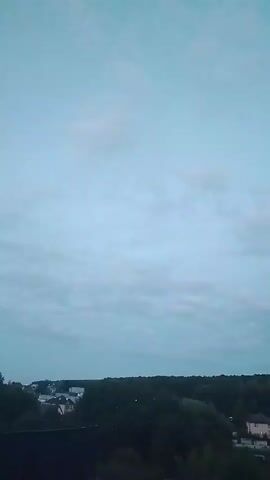 The video captures a panoramic view of a suburban landscape at dusk. The sky is a pale blue, with scattered clouds and a subtle gradient towards a lighter hue at the top. A dense line of dark green trees forms the horizon. Below the trees, several houses are visible, their lights beginning to illuminate as daylight fades. In the foreground, a dark, angled roof of a building suggests a house or other structure. The overall impression is one of a quiet, rural evening settling in.