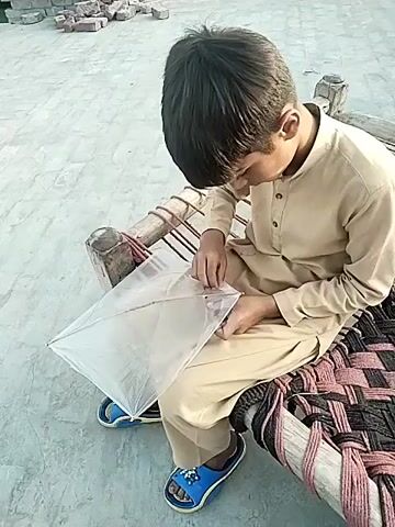 A boy sits on a bed with a woven seat, mending a transparent kite. The boy is wearing a light brown tunic and pants. He has short, dark hair and is focused on the kite. He is wearing blue sandals with decorations. The bed frame is wood, with rope interwoven for support. Behind him is a grey concrete surface with some scattered bricks. The lighting suggests it is daytime. There is a slight upward angle in the shot.