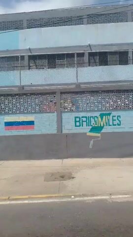 This video appears to be a shaky, first-person point-of-view recording taken from a vehicle moving along a street. The scene primarily depicts the exterior of a building with a light blue facade, multiple levels of windows with horizontal blinds, and a distinctive patterned brick wall. On the patterned wall, there is a mural. The first frame shows the word "BRICOMILES" in large, colorful block letters, with a paint roller icon below it. The mural transitions in the subsequent frames to a stylized, abstract red and yellow "M" shape. A portion of the Venezuelan flag is also visible on the wall.

Beyond the building, a chain-link fence encloses a parking area where a few vehicles are parked, including a white van and a dark-colored SUV. In the background, taller buildings and lush green mountains are visible under a bright, cloudy sky, suggesting it's daytime. The weather appears to be sunny but with scattered clouds. The presence of mountains and the architecture suggest a tropical or subtropical location, consistent with the provided context of Macuto, Venezuela. The overall mood is observational, likely capturing a passing view of the area. No people are clearly visible or interacting with the environment. The road in the foreground shows markings, indicating it's a public thoroughfare.