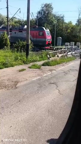 A train with red and gray cars travels along a track on a sunny day. The train is partially obscured by green bushes and trees. There is a road in the foreground of the video, and a power line pole stands between the road and the train tracks. The video is shot from a moving vehicle, likely a car, given the slight camera shake and the perspective. The overall environment appears to be rural or suburban, with a clear blue sky overhead.