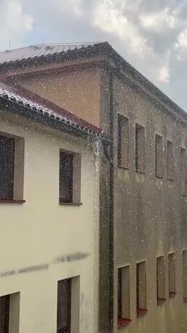 The video shows a close-up view of two adjacent buildings during a rainstorm. The building on the left is a lighter color, possibly beige or cream, with several windows visible, each framed in dark wood and with red window sills. The building on the right is a darker, yellowish-brown color and also has numerous windows with red sills, arranged in a regular pattern. A downspout or drainpipe is visible running vertically between the two buildings. Heavy rain is falling, creating a visible sheet of water cascading down the side of the building on the right and splashing onto the ground and grass below. Splashes and droplets of rain are also visible throughout the frame, obscuring some of the details of the buildings. The sky is overcast with clouds.