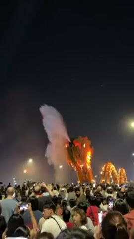 At night, a large crowd of people gathers on a bridge to watch a golden, illuminated dragon sculpture breathe a powerful stream of water into the air.  Many people are holding up their phones to record the spectacle. The sky is dark, and the dragon is the primary light source, along with streetlights.