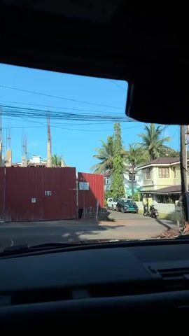 From inside a car, a view shows a residential street under a clear blue sky. The car is stationary. On the right side of the street, a two-story house painted in a pale yellow with brown accents is visible. A white picket fence surrounds the house, and there is a sign that reads "Dr. J.G. Noronha." Palm trees are scattered throughout the area. On the left side of the street, there's a red corrugated metal fence, likely indicating a construction site. A white car and a dark SUV are parked along the street, along with two motor scooters. The car's dashboard and navigation screen are visible in the foreground, showing a map.
