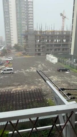 The video depicts a construction site viewed from above, likely from a balcony. The site features a partially constructed building with visible rebar and concrete. Several vehicles are present, including two white vans, a white SUV, and a black car parked. There are piles of construction materials and debris scattered around. The environment appears to be overcast with rain falling, evidenced by visible raindrops in the air and on surfaces. In the background, other buildings and trees can be seen. The camera appears to be mounted on a railing, indicating a vantage point from a residential building overlooking the construction area.