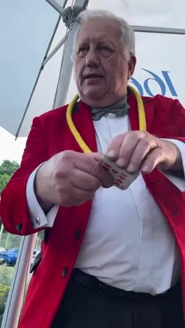 A man in a red jacket and a bow tie performs a magic trick with playing cards. He holds a card, the 10 of hearts, and appears to be manipulating it with his hands. The video is shot from the chest up, with the man's face and hands as the primary focus. He is outdoors, with a white tent or umbrella visible above him. There is a blue banner with white lettering in the background, though the text is not entirely clear. The man's expression is focused on his performance.