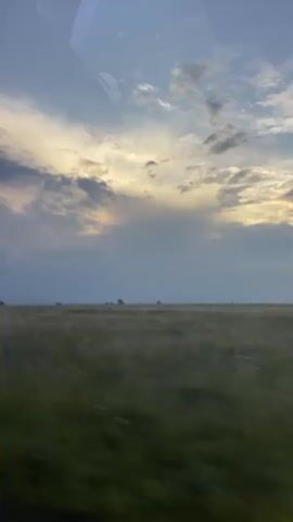 The video shows a wide, flat, grassy field under a cloudy sky at dusk. The camera appears to be moving, possibly from inside a vehicle, as indicated by the reflection in the upper portion of the frame and the slight motion blur in the foreground. The sky is a mix of blues, grays, and yellows, with sunlight filtering through some of the clouds. There are a few scattered trees and fence posts visible in the distance, suggesting a rural or open landscape. No people or animals are visible. The overall atmosphere is serene and slightly melancholic, characteristic of a late evening in an open area.