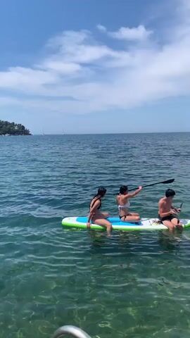 Three young people are on a paddleboard in the ocean. The person on the left is a female with dark hair, wearing a black bikini. She is facing the right and has her legs bent. The person in the middle is also a female with dark hair, wearing a white bikini. She is facing the right and is holding a paddle, appearing to paddle the board. The person on the right is a male with dark hair, wearing dark swim trunks. He is facing the left and is also holding a paddle. The paddleboard is white with green and blue accents. The water is clear and calm, with the seabed visible below. The sky is blue with some white clouds. There are boats visible in the distance. The video captures a sunny day at the beach.
