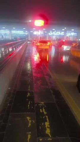 The video depicts an exterior scene at night, possibly at an airport parking area. It is raining, and the ground is wet and reflective. The camera moves along a walkway or raised platform, with a view of parked cars below. There is traffic moving through the area. The scene is lit with artificial lights, including red tail lights from vehicles and overhead streetlights. A large building can be seen in the background.