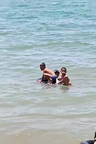 Three children are in the ocean, close to the shore. Initially, they are huddled together. Then, one child climbs into a dark-colored inner tube with pink and white stripes. Another child holds onto the tube while standing in the water. The water is relatively calm with small waves. The weather appears sunny and bright.