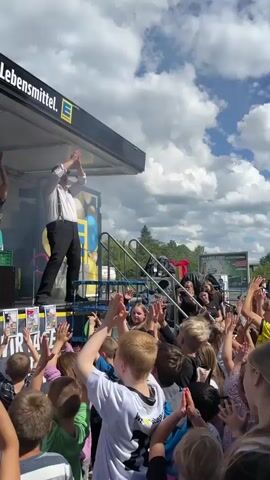 A group of people are on a stage, clapping their hands. They are all looking upwards and appear to be participating in some kind of performance or activity. There is a banner above them that reads "Wir lieben Lebensmittel" which translates to "We love groceries". Below the stage, a crowd of people, mostly children, are also clapping their hands and looking up at the stage. The scene appears to be an outdoor event, possibly a celebration or festival, given the bright lighting and the presence of a crowd. There are some crates and boxes on the stage, suggesting props or part of the performance setup. The atmosphere seems lively and engaging.
