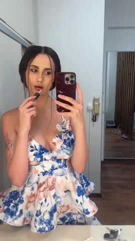 A young woman with dark hair tied up is applying lipstick in a selfie. She is wearing a strapless dress with a floral pattern of blue, pink, and white flowers. She is holding a dark red smartphone in her right hand and a lipstick in her left hand, applying it to her lips. Her gaze is directed towards the phone's screen. The background suggests an indoor setting, possibly a dressing room or a bathroom, with a mirror reflecting part of the room and a wooden slatted wall. There is a white sink or vanity in the foreground with some items on it, including keys and a small white object. The lighting appears to be artificial, suggesting it is not daytime. There is no visible text in the media.