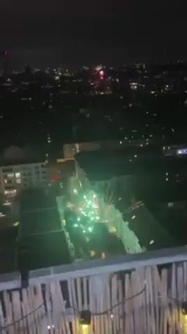 The video shows a nighttime cityscape with fireworks. The fireworks explode at a lower altitude than the distant city skyline, appearing closer to the buildings in the immediate foreground. The camera shakes during the explosions. The fireworks are a mix of colors, including orange, yellow, and red. The immediate surroundings appear to be buildings with some visible lighting. The video appears to be taken from a balcony or elevated position.